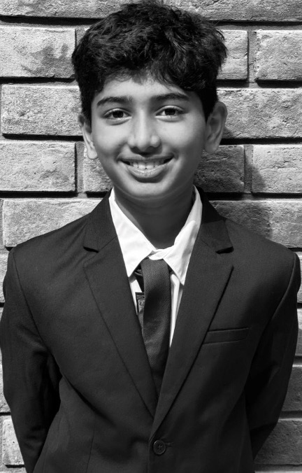 Portrait of a smiling student in a dark blazer, white shirt and tie, standing in front of a brick wall.