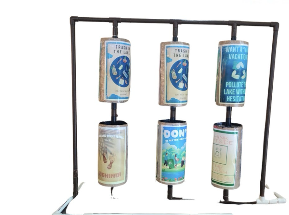 “The Rollers” installation: a dark pipe frame holding six rotating cylinders, each wrapped in a colourful anti-littering poster with slogans such as “Trash in the lake? Oh! What a great mistake”, “Leave nothing behind!” and “Think outside the trash”.